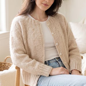 Könnte beinhalten: Eine beigefarbene Strickjacke mit Rundhalsausschnitt, langen Ärmeln mit gerippten Bündchen und einer Knopfleiste mit kleinen weißen Knöpfen. Das Strickmuster zeigt dezente vertikale Zopfmusterdetails entlang der vorderen Knopfleiste.