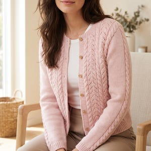 May include: A light pink knit cardigan featuring a cable knit pattern on the front panels and sleeves, a round neckline, and a button-down front with small, round, light-coloured wooden buttons. The garment is worn over a plain white t-shirt.