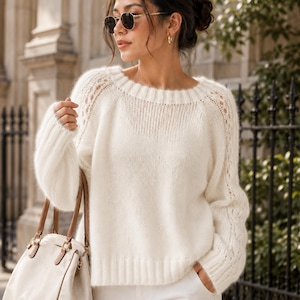 Puede incluir: Un suéter de punto de manga larga en color crema con cuello, puños y dobladillo acanalados, que presenta detalles de punto calado a lo largo de los hombros y las mangas, combinado con pantalones blancos y un bolso beige con asas de cuero marrón.
