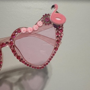 May include: Pink heart-shaped sunglasses with a flamingo accent on the upper right corner. The frames are adorned with pink rhinestones. The word "CLEO" is spelled out in pink letter beads across the left lens.