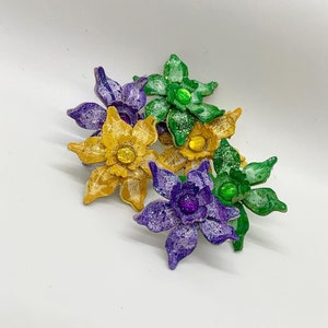 May include: A cluster of five glittery flower-shaped embellishments in shades of purple, yellow, and green. Each flower has a central jewel-like accent.