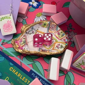 May include: A collection of game-themed accessories on a pink floral background. Includes a pair of pink dice, a decorative shell, and various game tiles. A dangling earring features the word "JOKER" and a playing card design.