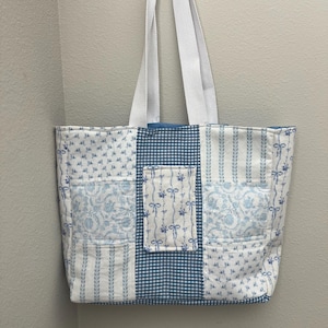 Puede incluir: Un bolso tote de patchwork con estampados florales, a rayas y de cuadros vichy en blanco y azul. El bolso tiene asas blancas y un pequeño bolsillo con un diseño de lazo. Diseñado para transportar objetos.