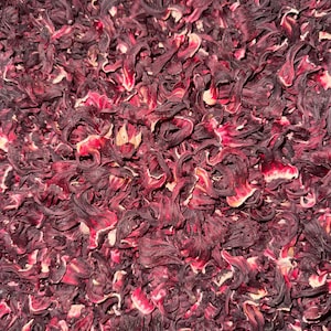 Op de afbeelding: Close-up van gedroogde hibiscusbloemen, met dieprode en bordeauxrode bloemblaadjes met lichtere roze en crèmekleurige accenten. De textuur is dicht en gelaagd, met een natuurlijke, organische uitstraling. De afbeelding suggereert kruidenproducten of culinaire ingrediënten.