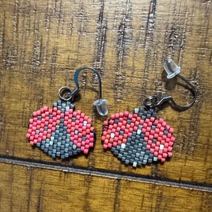 May include: A pair of handmade beaded earrings. Each earring is a ladybug design, with red, grey, and white beads. The earrings have silver-coloured ear wires and clear plastic earring backs. The earrings are displayed on a wooden surface.