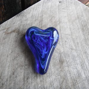 May include: A heart-shaped glass paperweight in shades of deep blue and white. The glass has a swirling pattern, creating a unique and artistic design. The heart is displayed on a light-colored, textured surface.