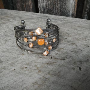 May include: A silver-toned cuff bracelet with multiple wire bands. The bracelet is adorned with various orange and peach-colored gemstones, including round and square shapes. The bracelet is displayed on a weathered wooden surface.