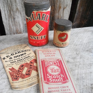May include: An assortment of vintage snuff tins and packaging. The largest tin is red with "SQUARE SNUFF" printed on it. Other items include a small tin with a bird graphic and paper packaging with "HONEST SCOTCH SNUFF" and "W.E. GARRETT & SONS" printed on them.