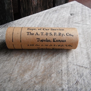 May include: A cylindrical, tan paper tube with black text. The text reads "Supt. of Car Service The A. T. & S. F. Ry. Co., Topeka, Kansas." The tube is resting on a weathered, light-colored surface.