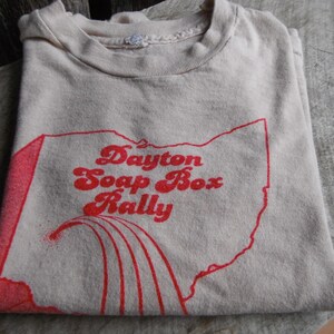 May include: A folded, beige t-shirt featuring a red graphic of the state of Ohio with the words "Dayton Soap Box Rally" in a retro font. The shirt is made of a soft, worn material, suggesting a vintage item.