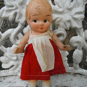 May include: A vintage doll with a painted face, wearing a red dress, white bib, and black shoes. The doll has a bald head and is posed on a white, ornate bench. The doll's arms are outstretched.