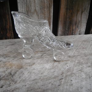 May include: A clear pressed glass figurine shaped like a high-heeled shoe with a starburst pattern across the surface. The shoe features a raised heel and a pointed toe, with a hollow interior and a smooth, transparent finish throughout the piece.