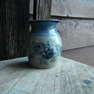 May include: A small, ceramic vase with a blue rim and floral design. The vase features a light beige base with blue floral patterns. The top and rim are a darker shade of blue. The vase is sitting on a light-colored wooden surface.