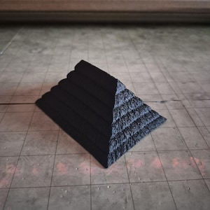 May include: A black, stepped pyramid-shaped object. The pyramid has a textured surface and appears to be made of a solid material. The pyramid is sitting on a tiled surface.