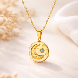 Peut inclure: Collier doré avec un pendentif représentant une lune en croissant et une étoile. Le pendentif comporte une pierre précieuse bleu clair au centre de l'étoile. Le collier est sur fond blanc.