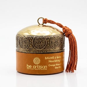 May include: A cylindrical lip balm container with a tan leather-like base and a metallic gold lid featuring an embossed geometric pattern. A rust-colored tassel hangs from a ring on top. The text reads be artisan, BAUME à lèvres, Nourishing Lip balm.