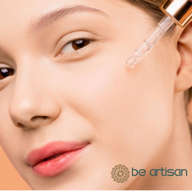 Pu&ograve; includere: Primo piano del viso di una persona con un contagocce che applica un liquido trasparente. La persona ha la pelle chiara e le labbra rosa. Il contagocce &egrave; tenuto vicino alla guancia. L'immagine include il testo "be artisan" e un logo decorativo.
