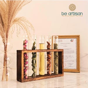Pu&ograve; includere: Un espositore in legno con cinque provette di vetro contenenti vari prodotti botanici e liquidi. Le provette sono disposte in un supporto di legno marrone scuro. L'immagine include anche un certificato incorniciato e il testo "be artisan moroccan beauty heritage".