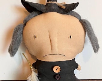 May include: A stuffed fabric doll shaped like a witch with a beige face, gray yarn hair, and a black pointed hat. The doll wears a black dress adorned with three round buttons in tan, dark brown, and red, and holds a small broom made of straw.
