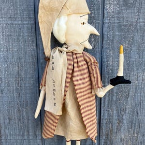 May include: A handmade doll with a long nose and a tall hat. The doll wears a beige dress and a red and beige striped coat. A scarf around the neck reads "bah humbug". The doll holds a candle with a black base and a yellow flame.