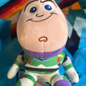 May include: A plush Buzz Lightyear doll, featuring a smiling face with embroidered details. The doll has a light pink head, blue eyes, and a green, white, and purple spacesuit. The background is a colorful patterned blanket.