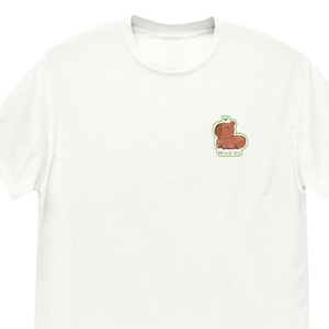 Peut inclure: T-shirt blanc avec un petit ours brun de dessin animé sur la poitrine. L'ours a un contour vert et un cœur au-dessus de sa tête, avec le mot "REEDING" en dessous. Le t-shirt a un col rond et des manches courtes.