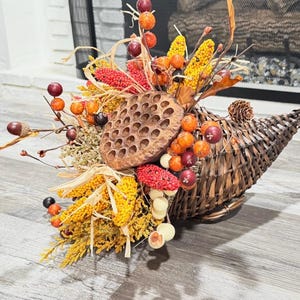 May include: A woven dark brown cornucopia basket filled with autumn-themed decor, including a large brown lotus seed pod, yellow millet stalks, red berries, orange berries, small pinecones, acorns, and dried grasses in shades of gold and tan.