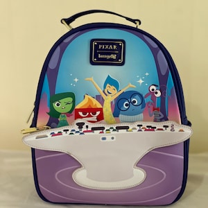 Può includere: Uno zaino viola e blu con i personaggi del film Pixar "Inside Out". Lo zaino ha una maniglia superiore, una tasca frontale e una chiusura a cerniera. Il design include Gioia, Tristezza, Rabbia, Paura e Disgusto. Il marchio "Pixar Loungefly" è visualizzato.
