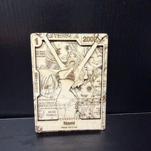 Puede incluir: Tarjeta de madera cortada con láser que presenta al personaje Nami de la tripulación del Sombrero de Paja. La tarjeta tiene un diseño detallado con múltiples imágenes y texto, incluyendo los números "1" y "2000".