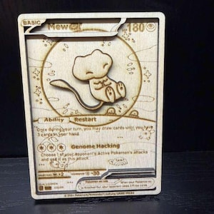 May include: A laser-cut wooden card depicting the Pokémon Mew. The card features detailed engraving of the character and surrounding design elements. Text includes "BASIC", "Mew", "HP 180", and ability descriptions.