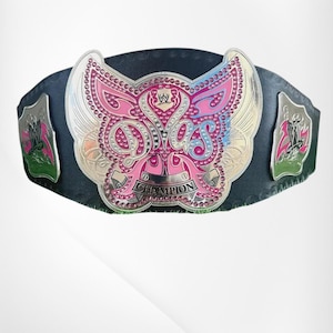 May include: A black leather championship belt featuring a large, butterfly-shaped silver metal center plate. The plate is decorated with pink and blue accents, embedded rhinestones, and the text Divas Champion with the WWE logo at the top.
