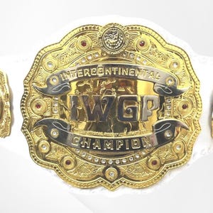 Può includere: Una cintura da campionato di wrestling dorata con una grande placca centrale recante il testo IWGP INTERCONTINENTAL CHAMPION, un emblema di leone in alto e piccole pietre rosse e trasparenti incastonate nella cornice metallica su cinturino bianco.