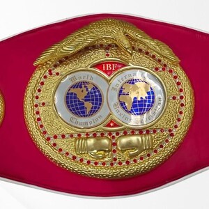May include: A championship boxing belt featuring a vibrant pink strap and a large central gold-tone plate. The plate displays an eagle emblem, two globes, the text IBF World International Boxing Federation, and two gold boxing glove icons.