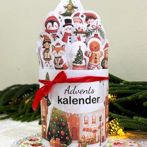 May include: A cylindrical advent calendar featuring a red ribbon tied around the top. The exterior displays a festive town scene with a Christmas tree and gifts. The top edge is lined with various die-cut holiday characters and icons.