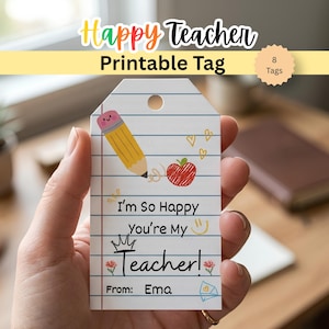 Può includere: Un'etichetta rettangolare in carta bianca con linee orizzontali blu e un margine verticale rosso. Presenta una matita, una mela rossa, piccoli cuori e il testo: I'm So Happy You're My Teacher! From: Ema. È presente un foro nella parte superiore.