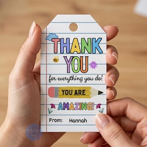 Op de afbeelding: Een wit cadeaulabel met een ontwerp van gelijnd papier, vastgehouden door een persoon. Het label bevat kleurrijke tekst met de tekst "THANK YOU", "YOU ARE AMAZING!" en "for everything you do!". Een potloodafbeelding en de naam "Hannah" zijn ook opgenomen.