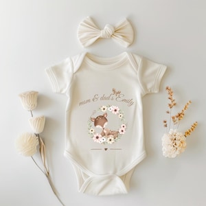 Puede incluir: Body de bebé color crema con un diseño de ciervo y corona floral. El texto dice "mom & dad's Emily". Un lazo color crema a juego está encima. Flores decorativas a la izquierda y a la derecha.