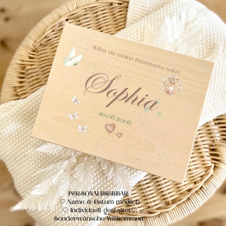 May include: A light-coloured wooden box featuring the name "Sophia" in a decorative font. The date "20.06.2006" is below the name, along with two hearts. The box is decorated with butterflies and the text "Willst du meine Patentante sein?"