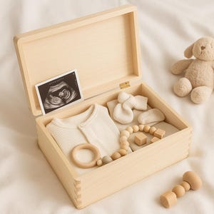 May include: A light wooden box, open to reveal baby essentials. Inside are a cream-coloured onesie, socks, a wooden teether, and a beaded toy. An ultrasound scan and a soft toy are also visible.