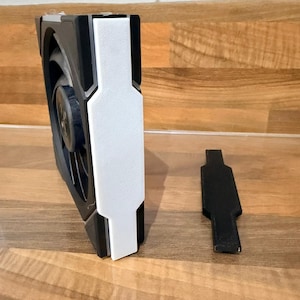 May include: A black and white computer fan with a white accent panel and a separate black component. The fan has a circular design with visible blades. The components are on a wooden surface.
