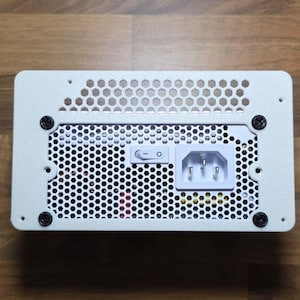 May include: A white power supply unit with a perforated top and a grid-like pattern. It features a power switch, an AC power inlet, and is secured with black screws. The unit is designed for electronic devices.