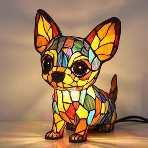 Könnte beinhalten: Eine Tiffany-Lampe in Form eines Chihuahua-Hundes, bestehend aus einem lebendigen Mosaik aus gelben, orangefarbenen, roten und blauen Glasstücken, die durch dunkle Metalllinien verbunden sind. Die Lampe hat große, dunkle, runde Augen und eine kleine schwarze Nase.