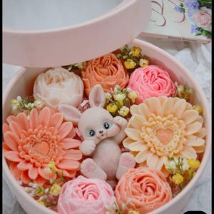May include: A circular, pale pink gift box containing soap flowers in peach, pink, and cream, encircling a small, light grey bunny figurine with blue eyes. The box is partly open, displaying the contents.