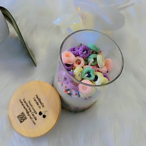 Op de afbeelding: Een glazen potkaars gevuld met kleurrijke, graanvormige stukjes was. De kaars heeft een houten deksel met tekst en een QR-code. Witte bloemen omringen de kaars, die op een zacht wit oppervlak staat.