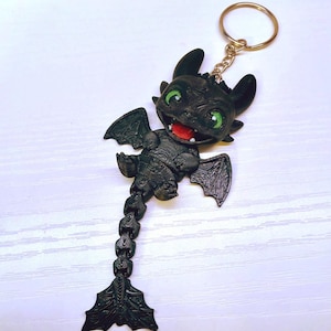 May include: A black dragon keychain with green eyes and a red mouth. The dragon has detailed wings and a tail, attached to a gold-colored keyring. The dragon is a stylized, three-dimensional design.