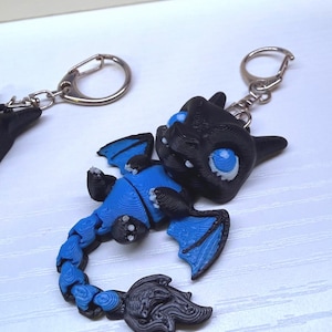 May include: A 3D-printed articulated dragon keychain featuring a black head, wings, and tail tip, with a vibrant blue body, wing membranes, and tail segments. The dragon has large, bright blue eyes and small white claws, attached to a silver metal keyring.