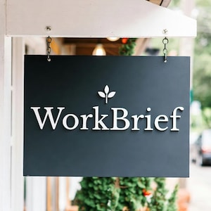 Könnte beinhalten: Ein rechteckiges, dunkelgraues Schild mit dem weißen Text "WorkBrief" und einem kleinen Blattdesign darüber. Das Schild ist mit Ketten an einer weißen Holzkonstruktion aufgehängt, im Hintergrund ist Grün zu sehen.