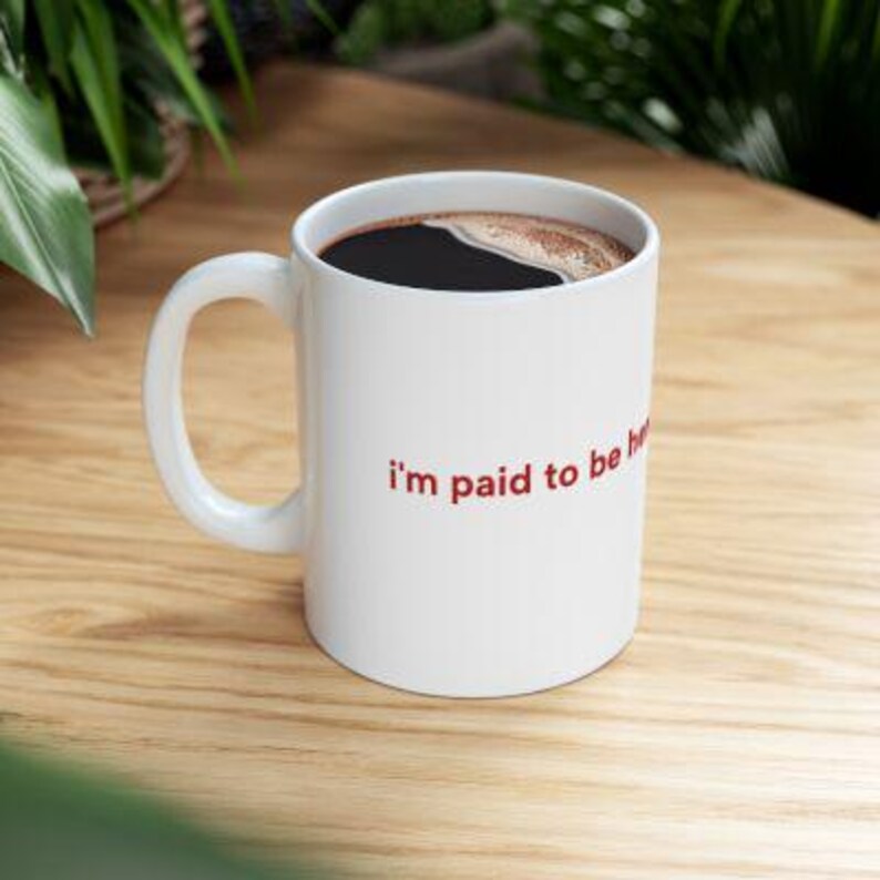 Puede incluir: Taza de caf&eacute; de cer&aacute;mica blanca llena de caf&eacute; oscuro. La taza tiene un asa curva y el texto "I'm paid to be here" en rojo. La taza est&aacute; sobre una superficie de madera clara.