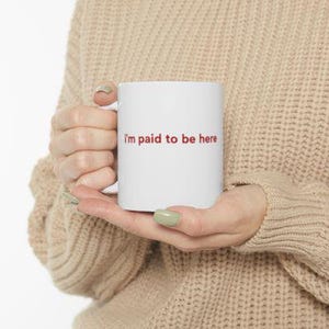 Puede incluir: Taza de cer&aacute;mica blanca con el texto "I'm paid to be here" en rojo. La taza la sostiene una persona que lleva un su&eacute;ter beige. La taza es de tama&ntilde;o est&aacute;ndar para caf&eacute; o t&eacute;.