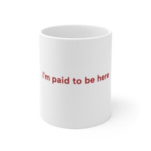 Puede incluir: Taza de cer&aacute;mica blanca con la frase "I'm paid to be here" impresa en rojo. La taza es cil&iacute;ndrica y tiene un dise&ntilde;o sencillo. Perfecta para caf&eacute;, t&eacute; o cualquier bebida.